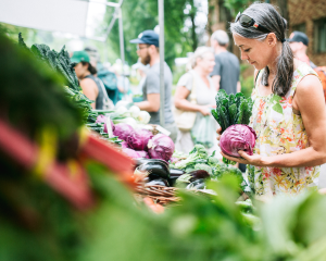 Outdoor produce stands at farmer's market with middle-aged woman in foreground holding purple cabbage