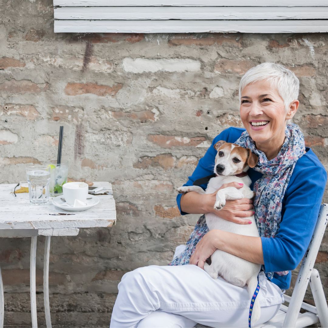Silver haired woman in bright blue jacket smiling while holder her Jack Russell terrier sitting at a cafe table outside