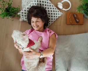older woman laying on floor with modern backdrop smiling at small white dog lying on her chest